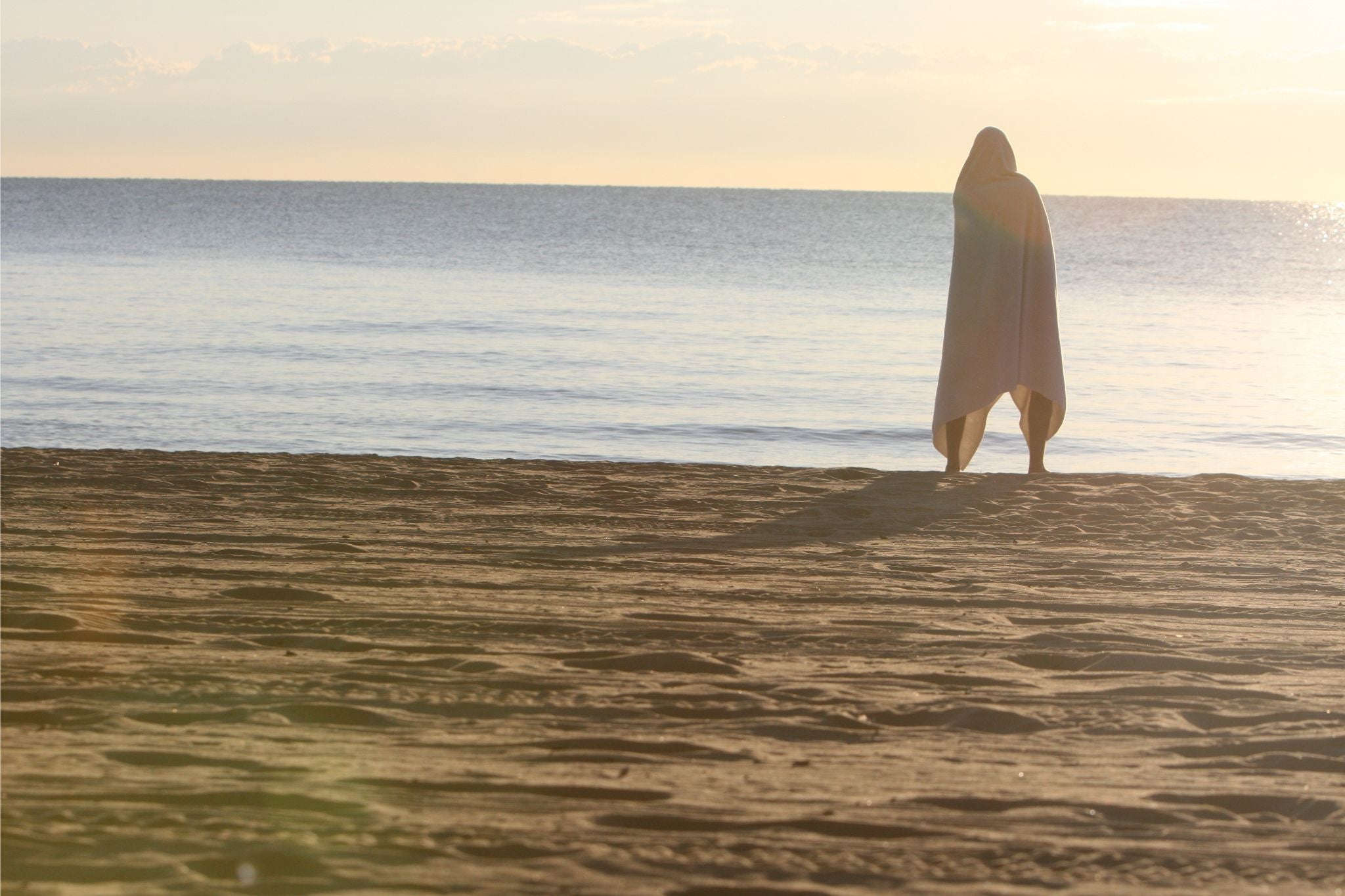 person wrapped in big towel overlooking the water by the beach
