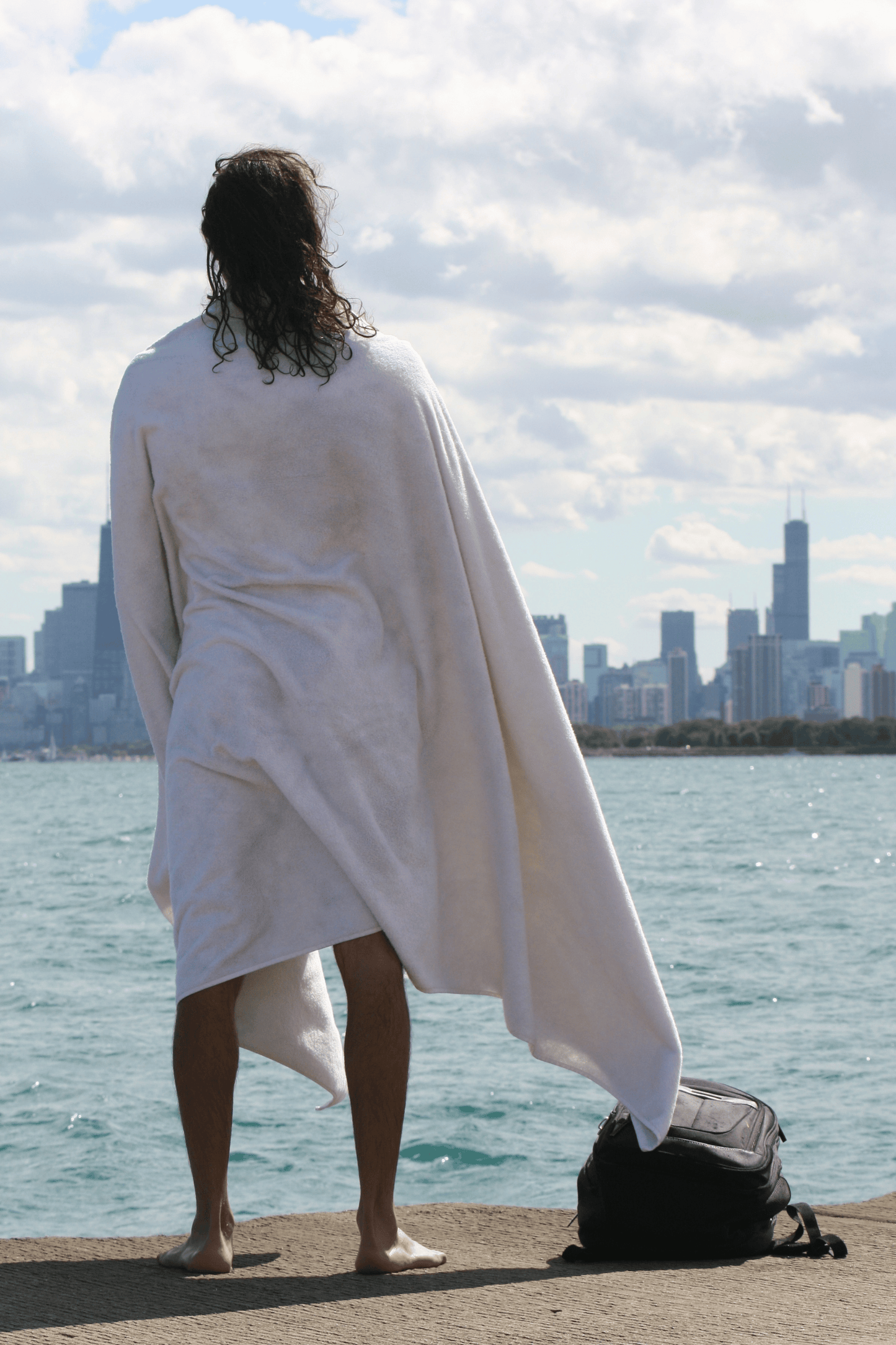 Person wrapped in a big white towel by a waterfront with a city skyline in the background
