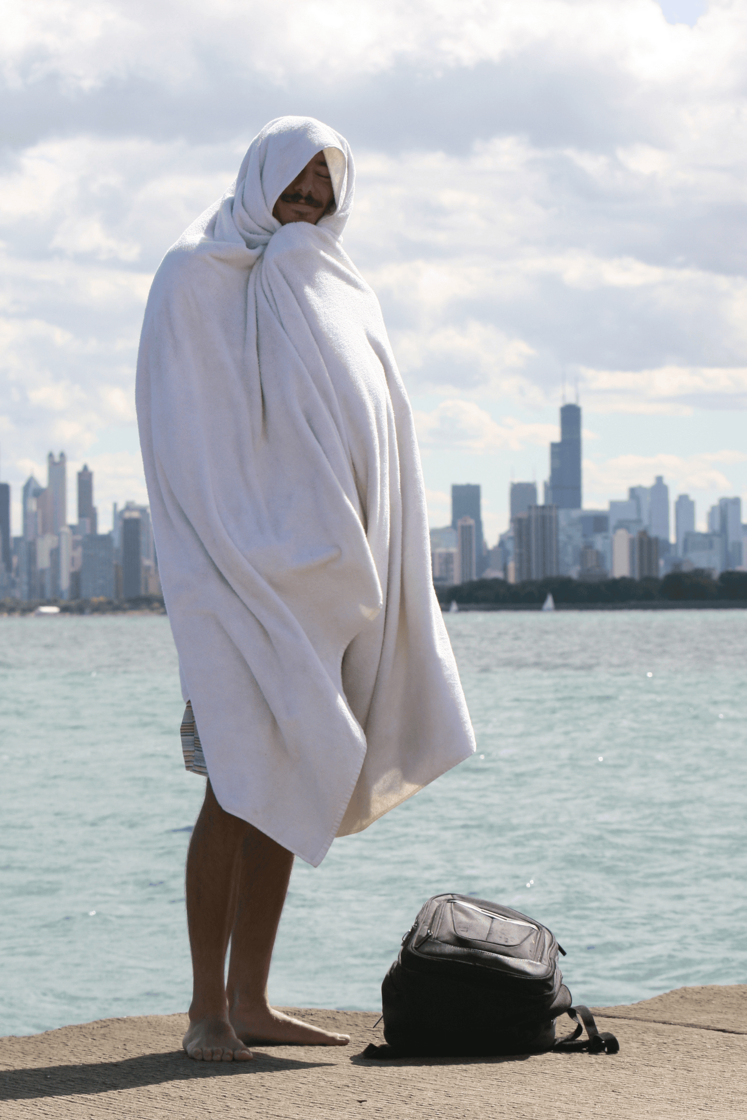 Person wrapped in a big towel standing by a waterfront with a city skyline in the background
